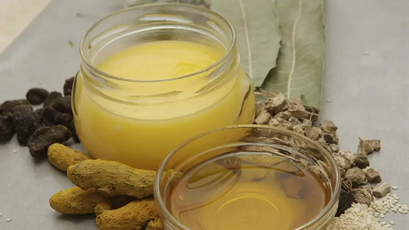 Pot de ghee, rhizomes de curcuma frais et bol d'huile sur fond ardoise, entourés de feuilles séchées