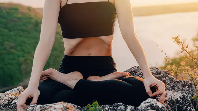 Femme assise jambes croisées, rétraction abdominale marquée, lors d'une pratique de Kapalabhati en extérieur au coucher de soleil