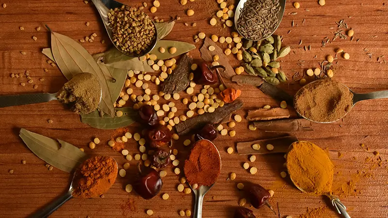 Vue plongeante sur huit cuillères d'épices ayurvédiques disposées en rosace autour de piments séchés et bâtons de cannelle, sur un plan de bois — curcuma, cumin, paprika, cardamome, laurier